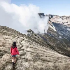 Une randonneuse explore un sentier herbeux, s'approchant des majestueuses montagnes du tour du Biros, enveloppées de nuages.