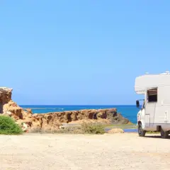 Camping-car blanc stationné sur une plage de sable en Espagne, avec la mer bleue et des falaises rocheuses en arrière-plan. Une aire de camping-car espagne gratuite idéale pour les vacances.