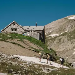 Trois randonneurs approchent d'une **cabane refuge** en pierre et bois, perchée sur une pente rocheuse et herbeuse, sous un ciel bleu intense.