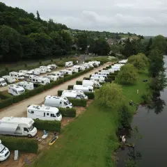 Vue aérienne d'une aire de camping-car gratuite en bord de mer normandie, avec de nombreux véhicules stationnés près d'une rivière bordée d'arbres.