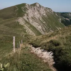 Sentier rocailleux serpentant dans l'herbe haute, menant vers des montagnes escarpées. Une clôture rudimentaire marque le chemin de cette échappée jurassienne.