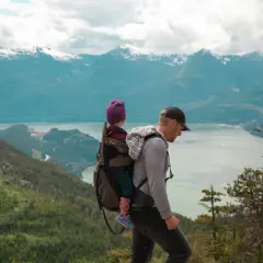Un père porte son enfant dans un porte-bébé lors d'un week-end randonnée, avec des montagnes enneigées en arrière-plan.