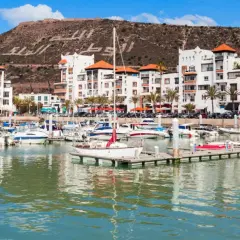 Port de plaisance à Agadir, Maroc, avec des bateaux blancs et des bâtiments modernes. La colline derrière porte une inscription en arabe.