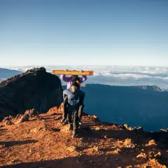 Couple joyeux au sommet du Piton des Neiges, portant une pancarte "Piton des Neiges 3070m". Paysage spectaculaire au lever du soleil.