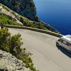Un camping-car blanc circule sur une route côtière sinueuse en Corse, dominant la mer d'un bleu profond.