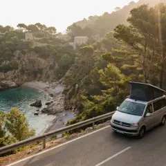 Un camping-car explore les routes sinueuses de la Costa Brava, offrant une vue imprenable sur une crique isolée et un phare au loin.