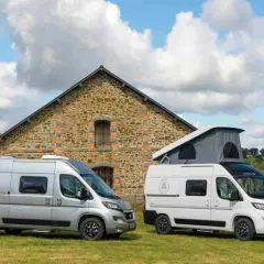 Deux camping-cars, un argenté et un blanc avec toit relevable, devant une bâtisse en pierre. Idéal pour savoir comment choisir son camping-car.
