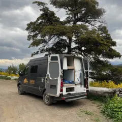 Un camping-car noir avec les portes arrière ouvertes, prêt pour un entretien, est garé près d'un lac sous un ciel nuageux.