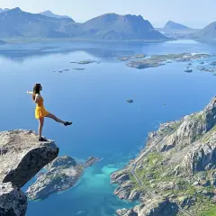 Une femme en robe jaune sur un promontoire rocheux, admirant les plus beaux endroits des îles Lofoten, avec des montagnes bleues et des eaux turquoise.