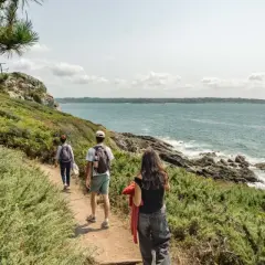 Trois randonneurs explorent le sentier des douaniers, longeant la côte rocheuse et la mer d'un bleu profond sous un ciel nuageux.