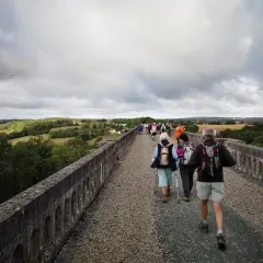 Un groupe de randonneurs, dont certains portent des sacs à dos et utilisent des bâtons de marche, traverse un ancien aqueduc en pierre sous un ciel couvert. Le paysage verdoyant s'étend à perte de vue.