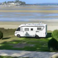 Vue d'une aire de camping-car avec sanitaire, face à une plage et un village côtier. Le camping-car blanc est garé sur l'herbe.