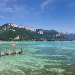Vue panoramique du lac d'Annecy, avec des montagnes verdoyantes en arrière-plan. Des bateaux et des pédalos animent les eaux turquoise. Un ponton fleuri invite à la promenade.