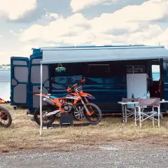 Un fourgon aménagé avec soute moto, prêt pour l'aventure. Deux motos de cross orange attendent devant, sous un ciel nuageux.