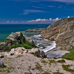 Sentier rocailleux menant au **Cap de la Chèvre**, avec falaises abruptes plongeant dans une mer turquoise et un ciel bleu éclatant.