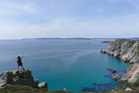 Randonneur victorieux au sommet d'un rocher, bras levés, dominant la mer turquoise et les falaises de GR34 Crozon.