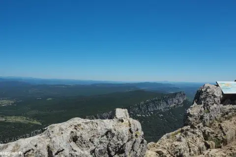 Vue panoramique depuis le pic Saint-Loup, avec une falaise imposante et une forêt verdoyante à perte de vue sous un ciel bleu éclatant.