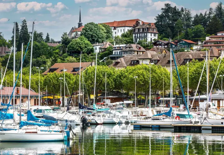 Port de plaisance à Thonon-les-Bains, avec des voiliers blancs et bleus amarrés, des maisons sur la colline et une église. Idéal pour savoir que faire à Thonon-les-Bains.