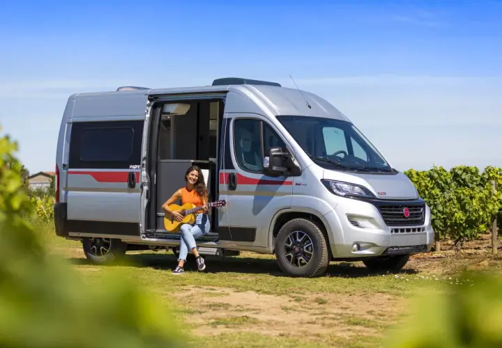 Une jeune femme joue de la guitare assise à l'entrée d'un camping-car aménagé, un type de camping car idéal pour les escapades ensoleillées.