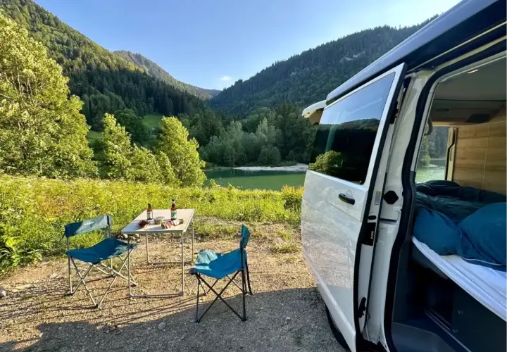 Un coin de paradis pour la Bourgogne en camping-car : table dressée, chaises bleues, lac paisible et montagnes verdoyantes.