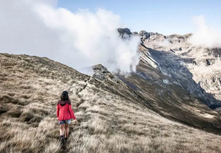 Une randonneuse explore un sentier herbeux, s'approchant des majestueuses montagnes du tour du Biros, enveloppées de nuages.