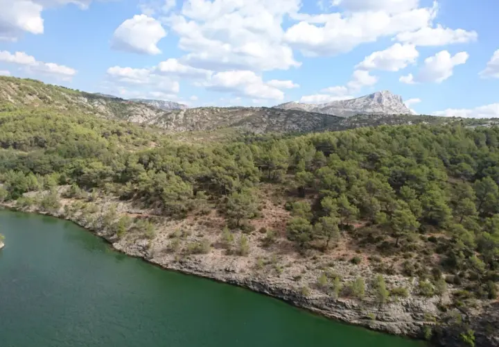 Vue panoramique du lac Zola, entouré de collines verdoyantes et rocheuses sous un ciel bleu parsemé de nuages blancs.