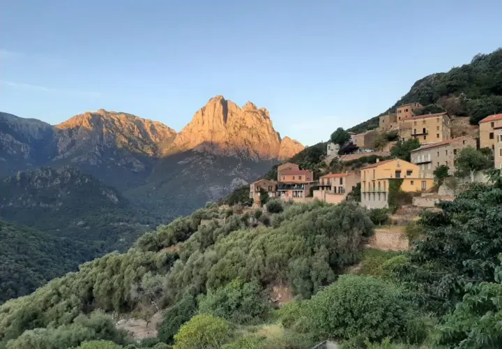 Village perché sur une colline, baigné par le soleil couchant. Les montagnes du nord, d'une difficulté certaine, se dressent en arrière-plan.