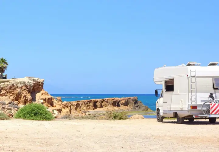 Camping-car blanc stationné sur une plage de sable en Espagne, avec la mer bleue et des falaises rocheuses en arrière-plan. Une aire de camping-car espagne gratuite idéale pour les vacances.