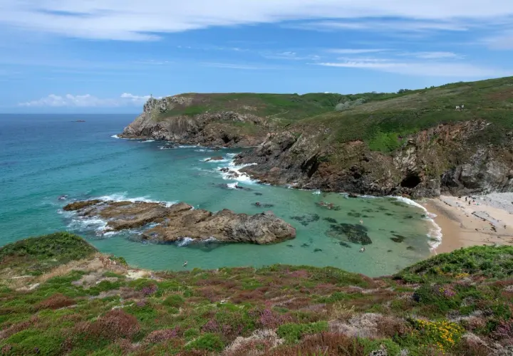 Vue panoramique de la pointe du Van, avec ses falaises verdoyantes, ses eaux turquoise et sa petite plage. Quelques baigneurs profitent du soleil.