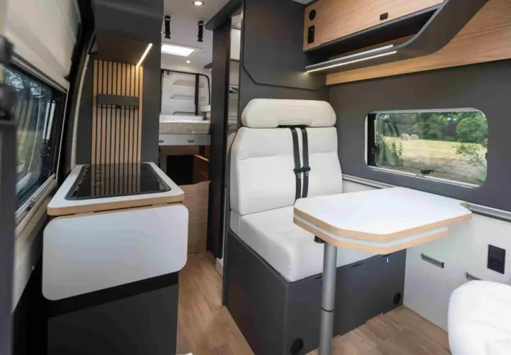 Intérieur d'un camping-car Bavaria : banquette confortable, table escamotable, plaque de cuisson et lit visible au fond.