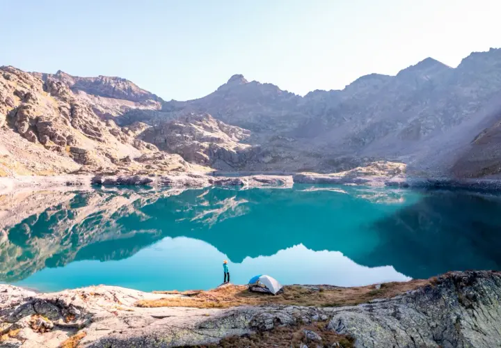 Un randonneur admire un lac turquoise aux reflets parfaits dans le **parc du Mercantour**. Une tente est installée sur la rive rocheuse.