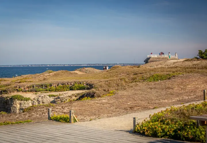 Vue panoramique sur la côte sauvage de la pointe Saint-Gildas, avec des falaises rocheuses, des dunes et la mer bleue. Des promeneurs profitent du paysage.