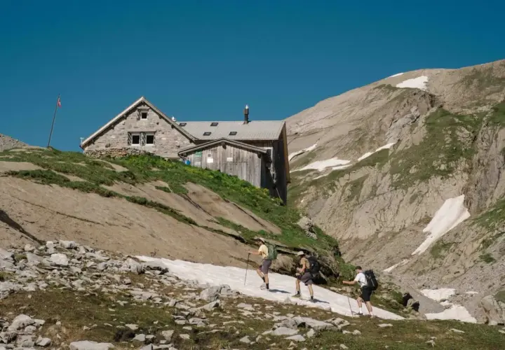 Trois randonneurs approchent d'une **cabane refuge** en pierre et bois, perchée sur une pente rocheuse et herbeuse, sous un ciel bleu intense.