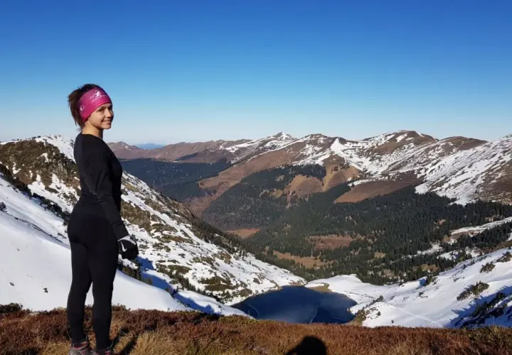 Vue panoramique depuis le pic du lion. Une femme souriante, en tenue de sport, admire le paysage montagneux enneigé et un lac gelé.