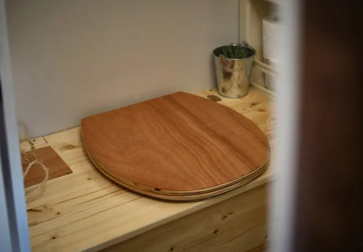 Un siège en bois pour toilettes sèches de camping-car, avec un petit seau de plantes et du papier toilette à proximité.