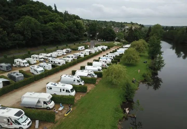 Vue aérienne d'une aire de camping-car gratuite en bord de mer normandie, avec de nombreux véhicules stationnés près d'une rivière bordée d'arbres.