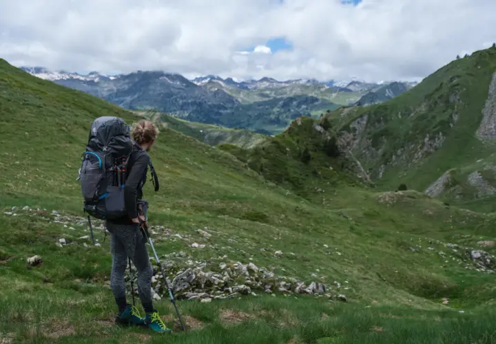Une randonneuse admire le panorama des Pyrénées, prête pour son trek. Le soleil perce les nuages sur les sommets enneigés.
