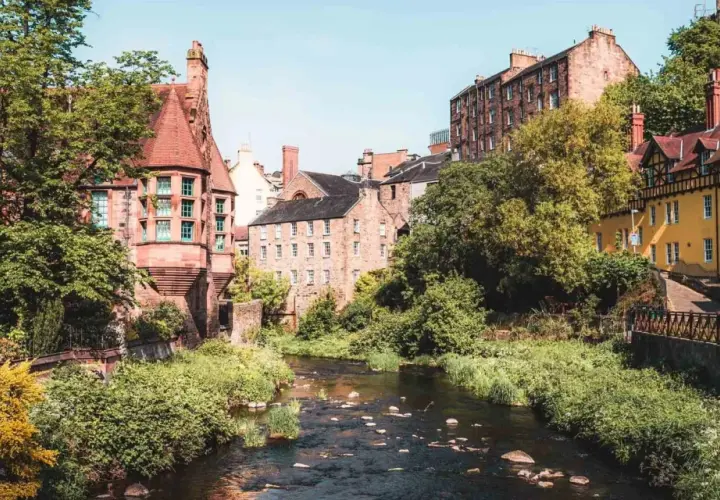 Un charmant ruisseau serpente à travers des bâtiments historiques en grès et une maison jaune vif. Idéal pour savoir que faire à Édimbourg.