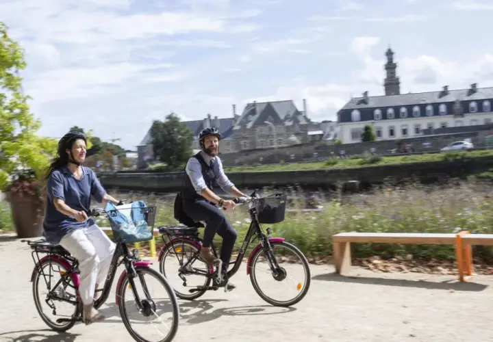 Couple à vélo profitant d'une balade ensoleillée le long de la rivière, avec les bâtiments historiques de Tréguier Lannion en arrière-plan.