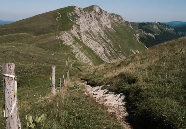 Sentier rocailleux serpentant dans l'herbe haute, menant vers des montagnes escarpées. Une clôture rudimentaire marque le chemin de cette échappée jurassienne.