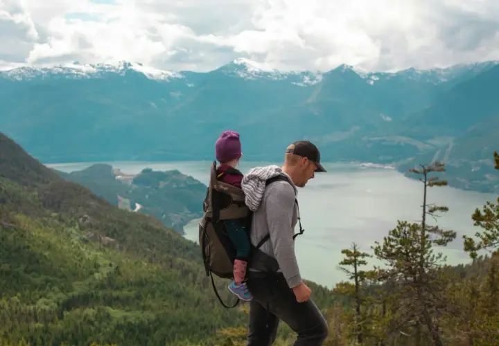 Un père porte son enfant dans un porte-bébé lors d'un week-end randonnée, avec des montagnes enneigées en arrière-plan.