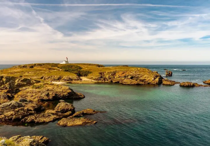 Phare blanc sur une colline verdoyante d'une île bretonne, avec des rochers dorés et une mer turquoise sous un ciel nuageux.
