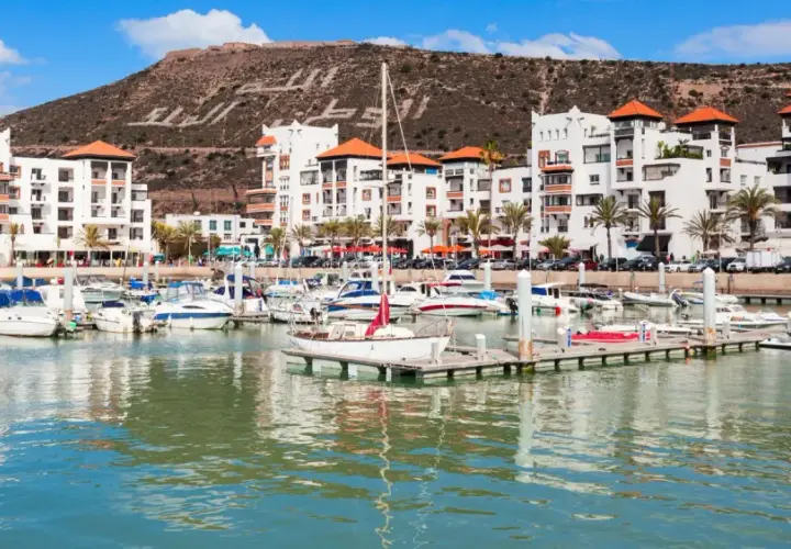 Port de plaisance à Agadir, Maroc, avec des bateaux blancs et des bâtiments modernes. La colline derrière porte une inscription en arabe.