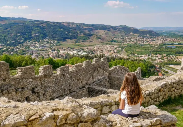 Une jeune fille contemple la vue depuis les ruines du **château de Crussol**, dominant une ville et des collines verdoyantes sous un ciel bleu.
