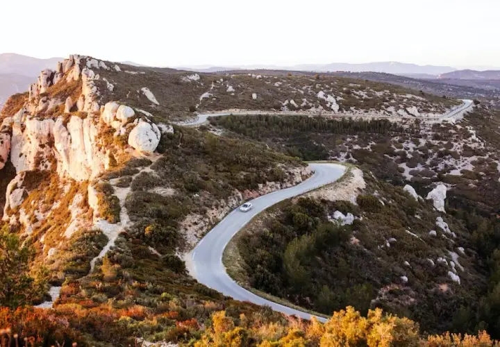 Route sinueuse à travers le massif du Cap Canaille, baignée par la lumière dorée du soleil couchant. Une voiture blanche serpente sur l'asphalte.
