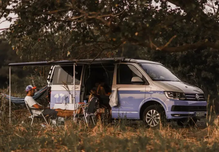 Un homme se détend près d'un camping-car sauvage, avec une table et des chaises installées à l'extérieur. Le véhicule est garé dans un cadre naturel verdoyant.