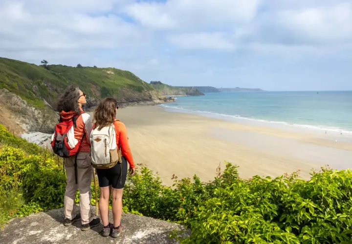 Deux randonneuses contemplent la plage de sable fin et la mer bleue, un paysage typique du GR34 à Saint-Brieuc.