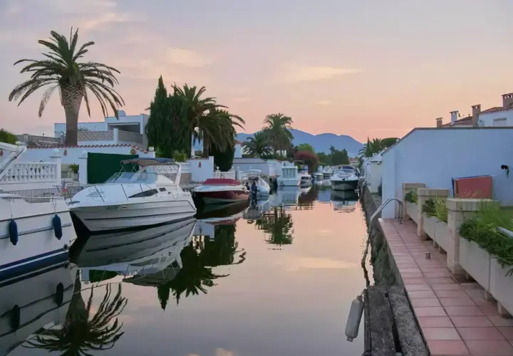 Coucher de soleil paisible sur les canaux d'Empuriabrava, Espagne, avec des bateaux amarrés et des palmiers se reflétant dans l'eau calme.
