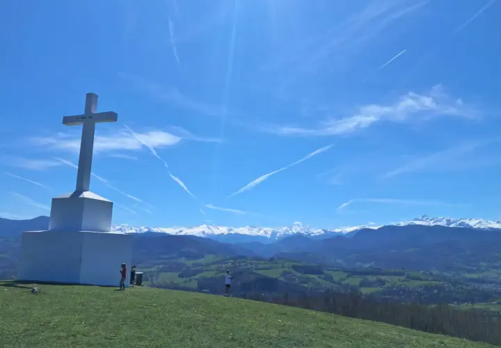 Le tuc de Montcalivert domine un paysage verdoyant sous un ciel bleu zébré de traînées de condensation. Une grande croix blanche se dresse fièrement.