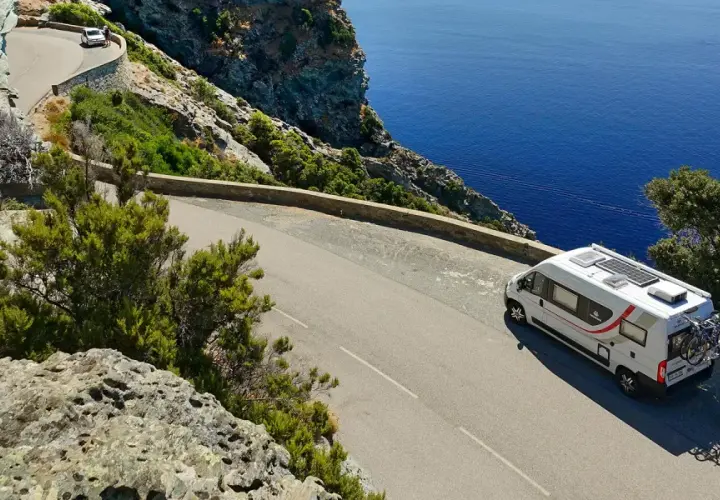 Un camping-car blanc circule sur une route côtière sinueuse en Corse, dominant la mer d'un bleu profond.